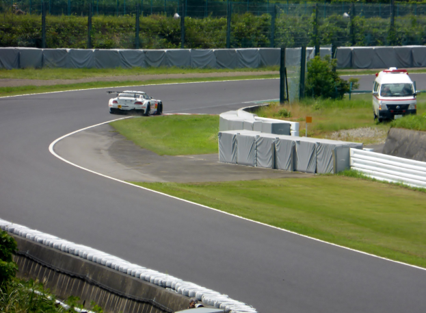 Ein Rennwagen, der auf einer Rennstrecke fährt, mit einem Polizeiauto im Hintergrund, umgeben von Gras, Pflanzen, Bäumen und einem Zaun, mit einem Fahrzeug auf der rechten Seite und Text am unteren Rand.