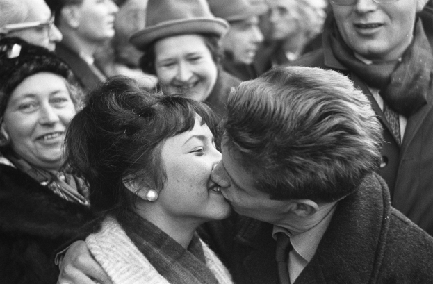 Ein Mann und eine Frau umarmen und küssen sich leidenschaftlich vor einer Menge lachender Menschen, dargestellt in Schwarz-Weiß.