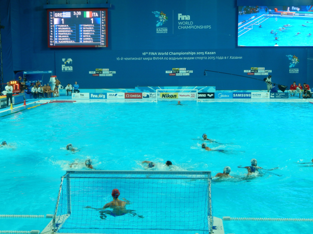 Wasserballspieler bei den 2016 FINA-Weltmeisterschaften in Kazan im Pool gegeneinander antretend, mit Zuschauern links und einem Anzeigebrett mit Event-Logo im Hintergrund.