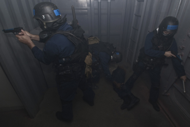 Eine Gruppe von Polizisten in Einsatzausrüstung steht zusammen, einer hält eine Waffe, mit einem Metallbehälter und einer Leiter im Hintergrund.