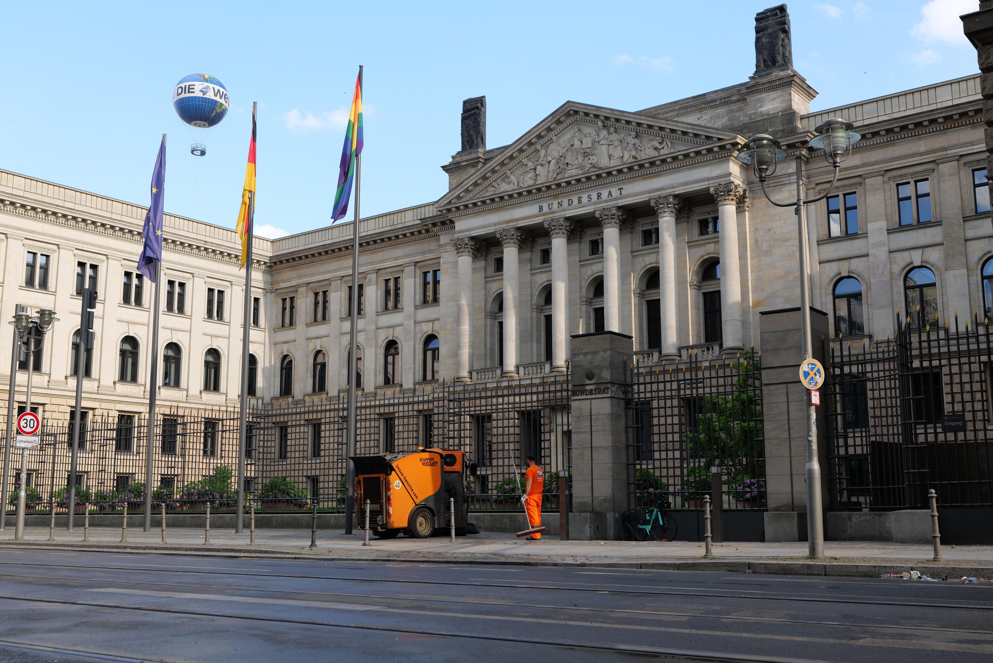 Großes beiges Gebäude mit vielen Fenstern, als Bundestag in Berlin, Deutschland identifiziert, umgeben von Straßeninfrastruktur, Fußgängern, Fahrzeugen und Bäumen unter einem bewölkten Himmel.