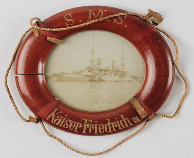 Roter Rettungsring mit 'USS Kaiser Friedrich III' Text und einem Bild eines Schlachtschiffs, an einer weißen Oberfläche mit Seilen befestigt.