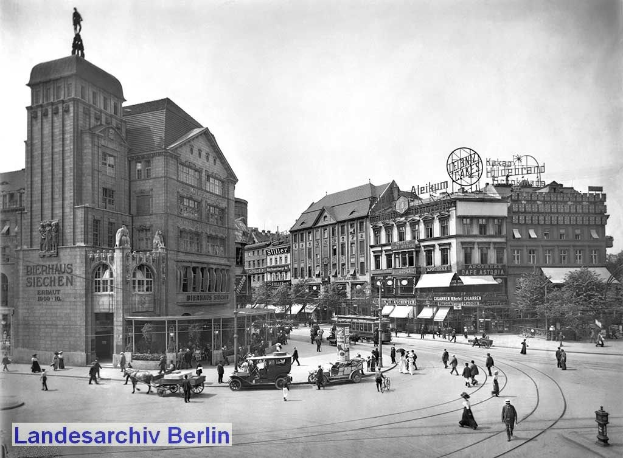Ein Schwarz-Weiß-Foto einer belebten Stadtstraße in Berlin, Deutschland, mit Menschen, Fahrzeugen, einer Statue an einem Gebäude, Pfählen, Schildern, Bäumen und einem bewölkten Himmel.