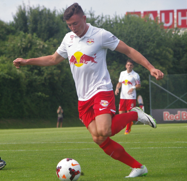 Fußballspieler bei einem Profispiel auf einem Rasenfeld mit "RB Leipzig"-Schriftzug im Hintergrund unter einem klaren blauen Himmel.