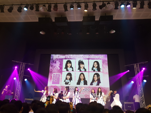 Eine K-Pop-Gruppe namens BK48 performs auf einer hell erleuchteten Bühne mit Performern, die ein Mikrofon halten und Instrumente spielen, während das Publikum unten aufnimmt und ein großer Bildschirm die Gruppenmitglieder anzeigt.