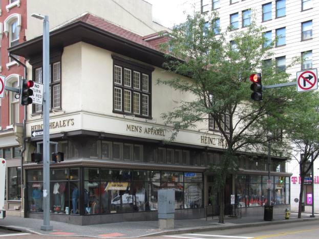 Eine belebte Straßenkreuzung mit einem geparkten Bus, Gebäuden, Bäumen, Laternen, Ampeln, Schildern und einem Kaufhaus mit Schaufensterpuppen im Hintergrund vor einem sichtbaren Himmel.