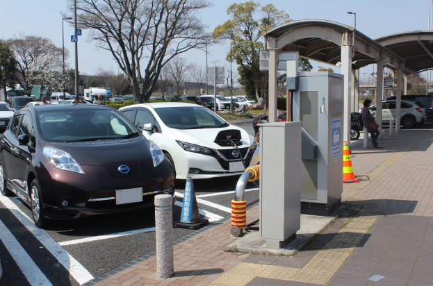 Elektroauto-Ladestation in Japan mit Autos auf der Straße, einer Person auf dem Gehweg und umgebender Infrastruktur wie Verkehrskegel, ein Schuppen, Mäste, Lichter und Bäume vor einem Himmelhintergrund.