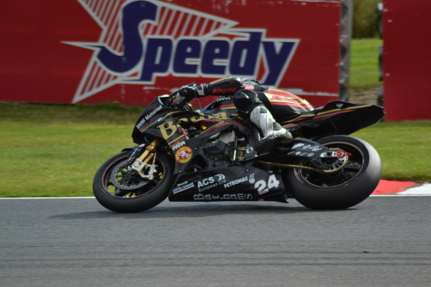 Eine Person in Helm und Schuhwerk fährt ein Motorrad auf einer Rennstrecke, umgeben von Gras, mit einer Tafel im Hintergrund.