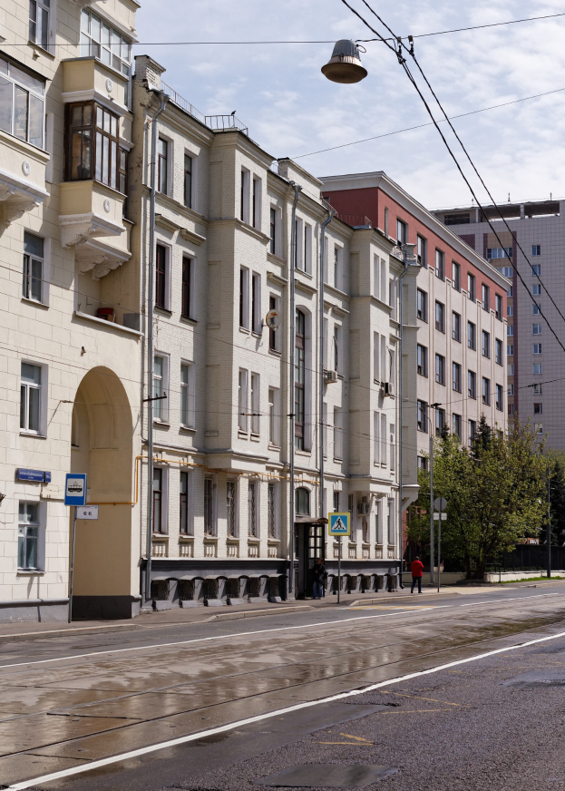 Eine nächtliche Stadtstraße mit hohen Gebäuden auf beiden Seiten, beleuchtet von Straßenlaternen, mit Strommasten, Drähten, Schildern, Bäumen und Fußgängern auf dem Gehweg, unter einem bewölkten Himmel.