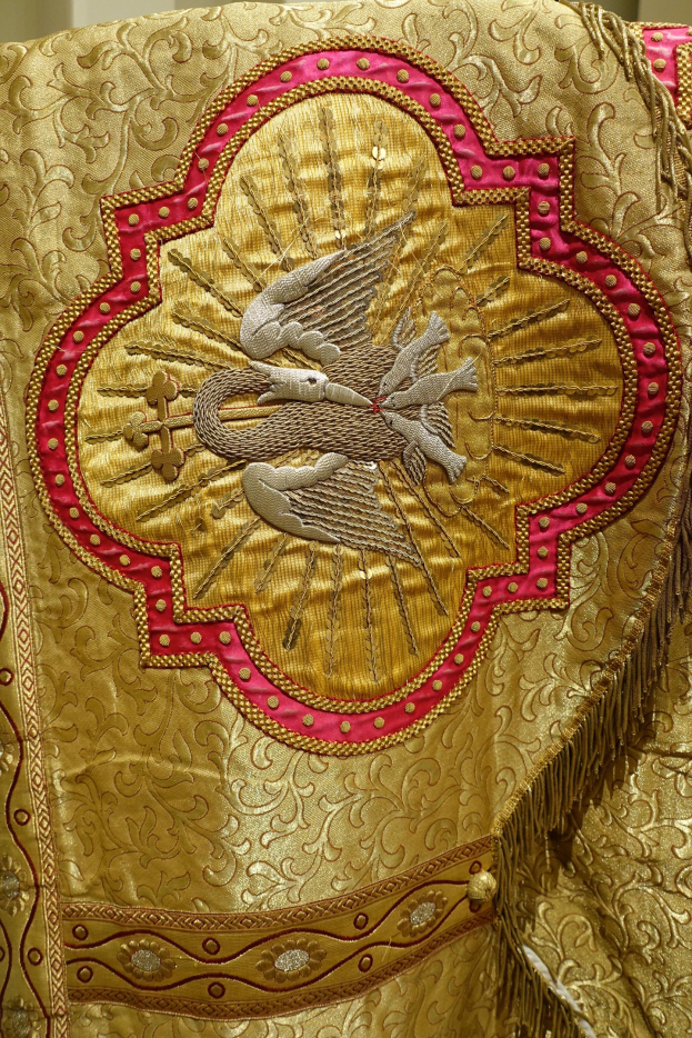 Nahaufnahme eines Priestergewandes mit goldenen und roten Stickereien und detaillierten Mustern vor einer Wand im Hintergrund.
