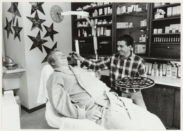 Ein Mann lächelt in einem Zahnarztstuhl, während ein anderer Mann daneben steht und ein Tablett hält, mit einem Waschbecken, Sternen, einer Uhr und Regalen voller Flaschen im Hintergrund.