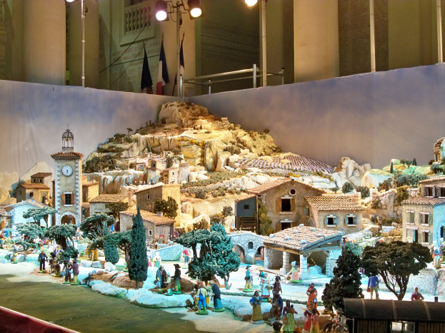 Eine Museumsausstellung einer Krippe mit Menschen, Häusern, Bäumen, Felsen, einem Kirchturm, Fahnen, Stangen und Lichtern, alles beleuchtet von Museumsbeleuchtung.