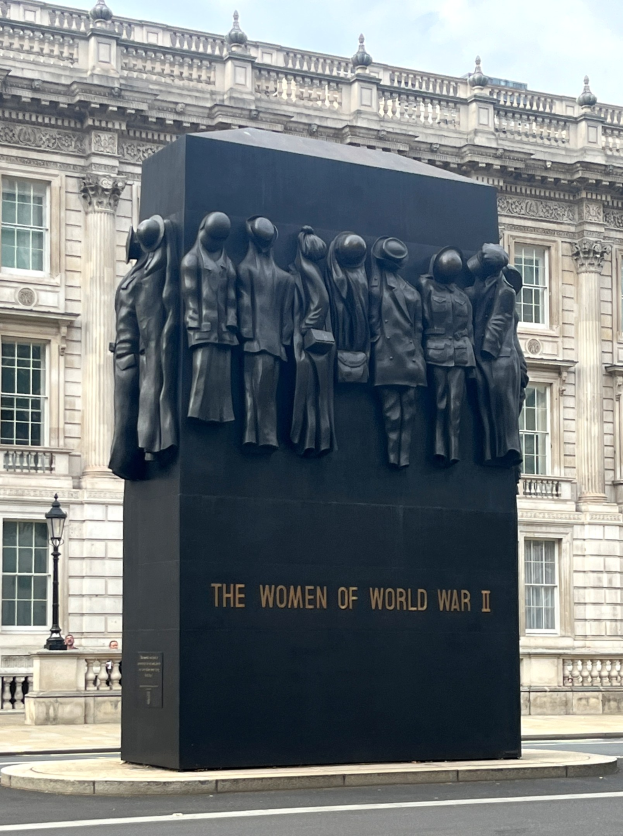 Das Frauen-Denkmal des Zweiten Weltkriegs in London, eine große schwarze Struktur mit eingravierten Text, umgeben von einem gepflasterten Bereich mit Geländern, Laternenmasten und einem Gebäude im Hintergrund unter einem bewölkten Himmel.
