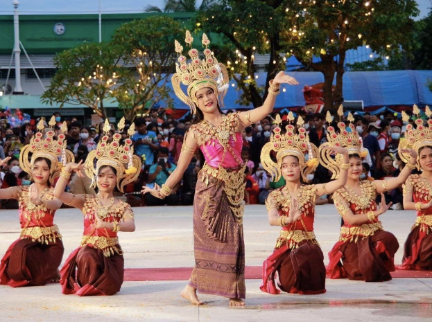 Gruppe von Frauen in traditioneller thailändischer Kleidung, die draußen tanzen, mit Zuschauern, Bäumen und einem Gebäude im Hintergrund bei klarem blauem Himmel.