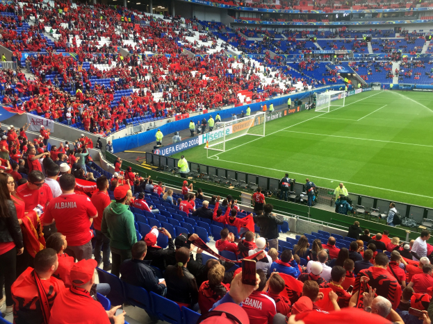 Eine große Menschenmenge in einem Stadion schaut einem Fußballspiel zu, mit Menschen, die auf Stühlen sitzen und stehen, Fahnen, einem Torpfosten und Deckenbeleuchtung.