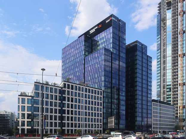 Großes modernes Bürogebäude mit vielen Fenstern, identifiziert als Deutsche Bank Hauptsitz in Frankfurt, Deutschland, umgeben von städtischer Infrastruktur einschließlich Straßenlaternen, Fahrzeugen, Bäumen und einem bewölkten Himmel.