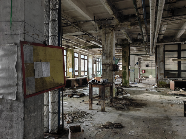 Verlassener Fabrikinnenraum mit Schutt, einschließlich Tischen, Stühlen und Papieren auf einer Tafel an der Wand, mit Fenstern, Säulen und freiliegenden Rohren im Hintergrund.