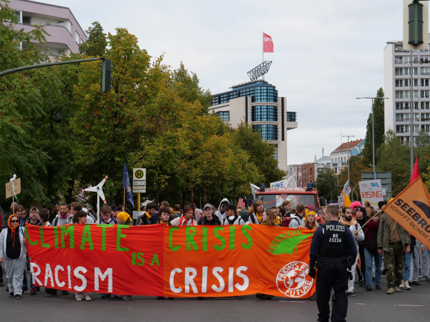 Eine Gruppe von Menschen marschiert auf einer von Bäumen gesäumten Straße, die ein Banner mit der Aufschrift "Klimakrise ist eine Krise" trägt, mit parkenden Fahrzeugen, Gebäuden und einem klaren blauen Himmel im Hintergrund.