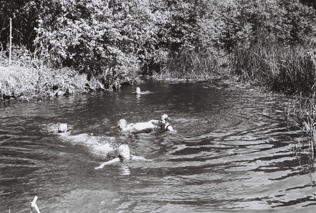 Schwarzes und weißes Bild von Menschen, die in einem Fluss umgeben von Bäumen und Pflanzen schwimmen.