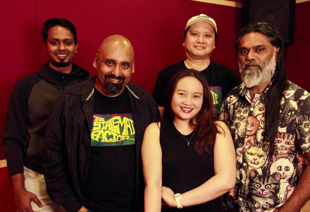 Eine Gruppe von Menschen in legerer Kleidung, die eng zusammenstehen und sich gegenseitig umarmen, lachend vor einer roten Wand in einem Aufnahmestudio mit einem Lautsprecher im Hintergrund.