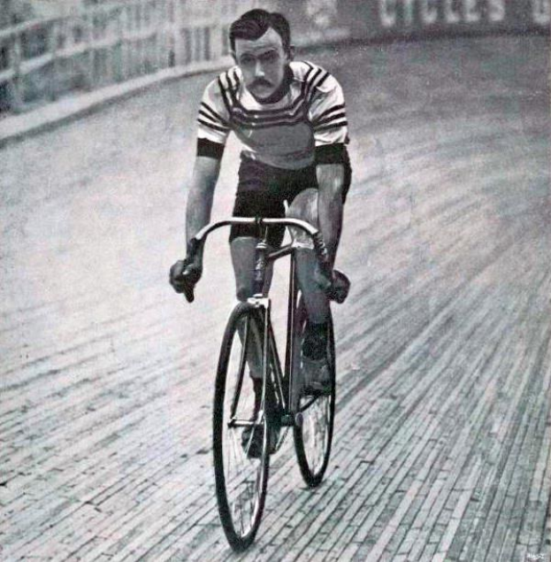 Schwarz-weiß-Foto eines Mannes, der auf einer Bahn radelt, mit einem Zaun und einer Tafel im Hintergrund.
