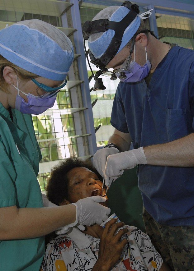 Zwei maskierte und handschuhgetragene Chirurgen führen eine Gesichtsoperation an einer Frau durch, die auf einem Bett liegt, mit Bäumen im Hintergrund durch ein Fenster sichtbar.