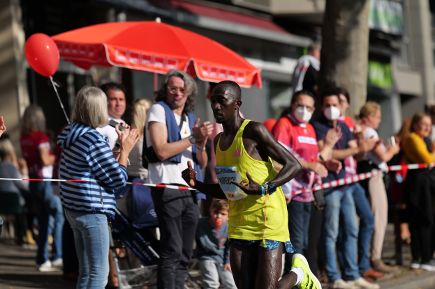 Ein Mann in einem gelben T-Shirt läuft an einer jubelnden Menge vorbei während eines Marathons, mit einem Schirm und einem Kinderwagen im Hintergrund.