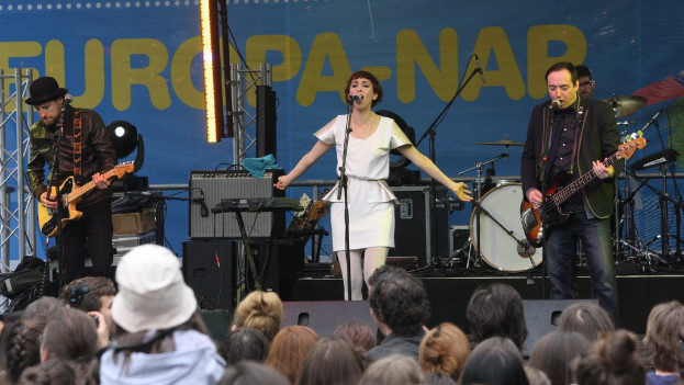 Vier Musiker treten auf der Bühne auf: Eine Frau in einem weißen Kleid singt in der Mitte, flankiert von zwei Männern, die Gitarren spielen, mit einer Menge unten und Lautsprechern an einer Wand im Hintergrund.