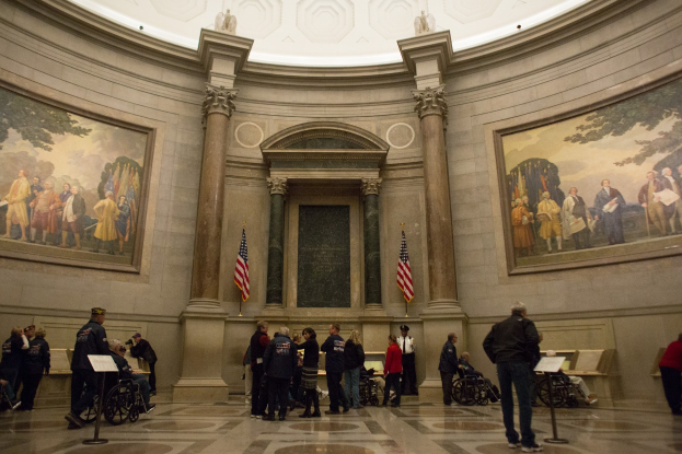Eine Gruppe von Menschen vor einem Gebäude mit Kuppel, einige im Rollstuhl und andere stehend, mit Ständen mit Papieren, Fotorahmen an den Wänden, Fahnenmasten und Deckenstatuen.