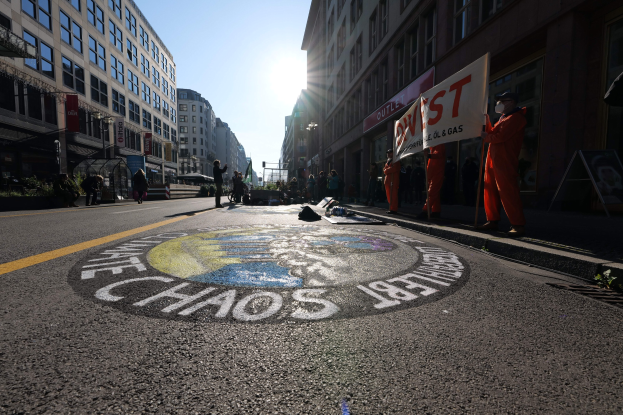 Eine Gruppe von Menschen hält Schilder und Plakate in den Händen und steht auf einer Straße, die mit Pflanzen und Gebäuden gesäumt ist, mit "Chaos" auf der Straße in Bezug auf die Occupy Wall Street Demonstration.