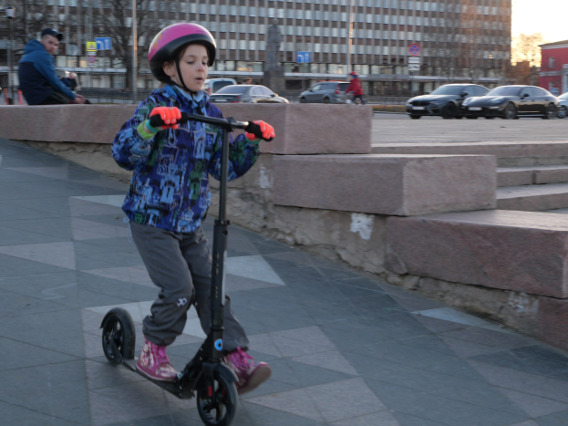 Ein junger Junge mit Helm und Handschuhen fährt auf einem Roller eine Treppe auf dem Gehweg hinunter, mit Fahrzeugen, Menschen, Bäumen, Polen, Brettern, Gebäuden und einem klaren blauen Himmel im Hintergrund.