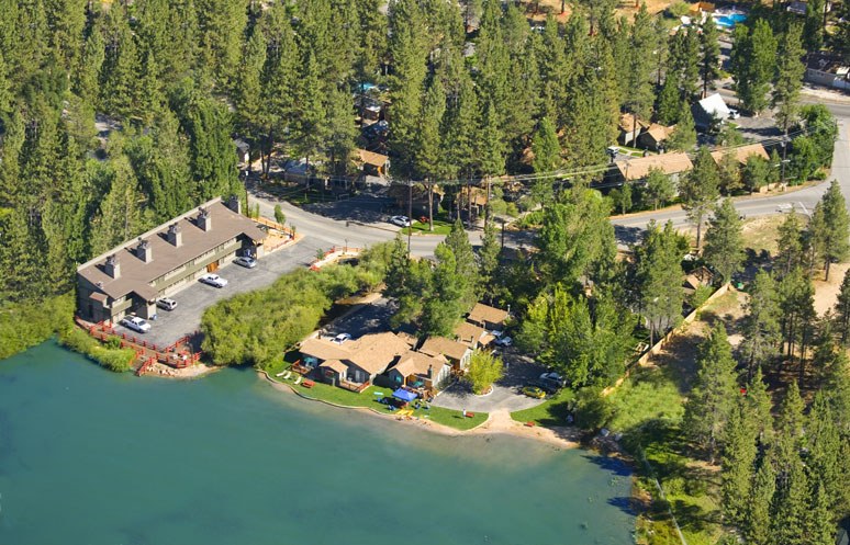 Luftaufnahme eines Resorts am Seeufer, umgeben von Baumen, Häusern, Fahrzeugen, Straßen, Masten und Gras, mit dem See unten.