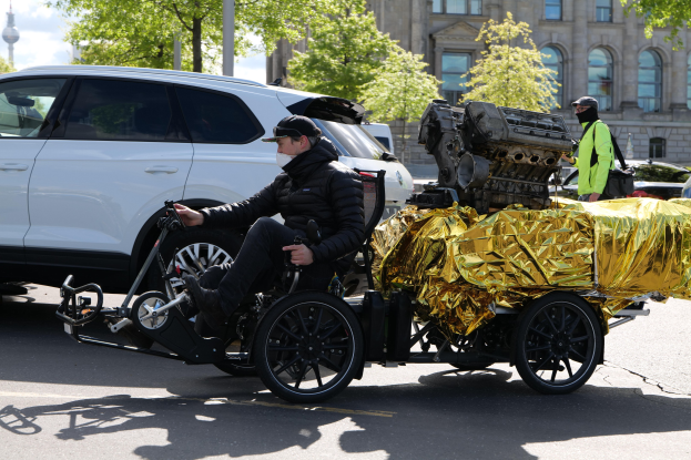 Ein Mann im Rollstuhl mit einem großen Motor an dessen Rðcken, umgeben von Fahrzeugen auf einer Straße mit Bäumen, Gebäuden und einem klaren blauen Himmel im Hintergrund; er trägt eine schwarze Jacke, eine Kappe und hält ein Objekt in der Hand.