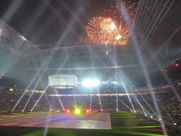 Das Olympische Stadion in London, England, nachts mit heller Beleuchtung, umgeben von einer großen Menge, grünem Gras und einem Himmel voller Feuerwerke.