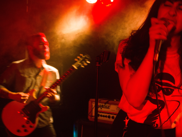 Eine Frau singt in ein Mikrofon auf der Bühne, während ein Mann im Hintergrund eine akustische Gitarre spielt, mit Deckenlampen, die die Szene beleuchten.
