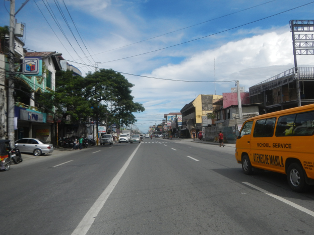 Ein gelber Schulbus fährt eine Straße mit hohen Gebäuden, Menschen auf dem Gehweg, Bäumen, Strommasten mit Drähten und Schildern entlang, unter einem bewölkten Himmel.