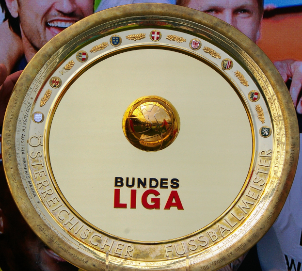 Ein Mann mit einem goldenen Schild, auf dem 'Bundesliga' steht, und einem Banner mit drei Personen im Hintergrund.