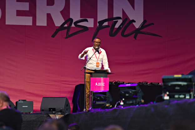 Ein Mann in einem weißen Hemd und braunen Hosen steht auf einer Bühne und hält ein Mikrofon in der Hand, hinter ihm eine pinkfarbene Fahne mit der Aufschrift 'Berlin as Fuck' und Lautsprecher auf der Bühne.