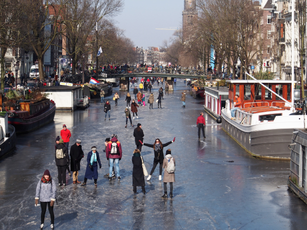Eine Gruppe von Menschen, die auf einem zugefrorenen Kanal in Amsterdam Schlittschuh laufen, umgeben von Booten, Bäumen, Gebäuden, Laternen, Flaggen und einer Brücke unter einem klaren Himmel.