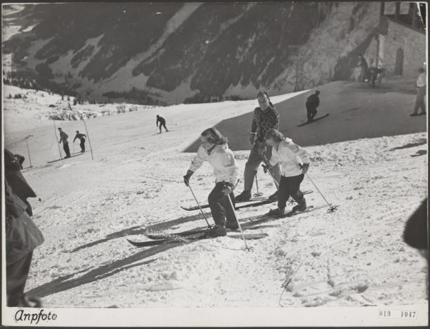 Gruppe von Menschen beim Skifahren auf einer schneebedeckten Piste mit Skistöcken, Hügeln und einem Gebäude im Hintergrund und Text unten.