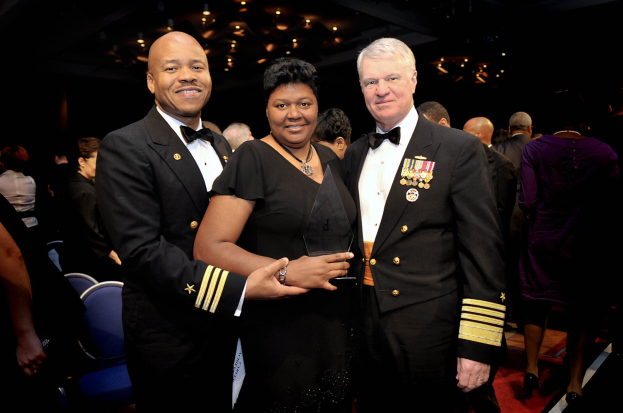 Zwei Männer in schwarzen Blazern und weißen Hemden stehen neben einer Frau in einem schwarzen Kleid, die eine Trophäe hält, auf einem roten Teppich, während andere im Hintergrund sitzen und stehen, unter beleuchteten Decken.