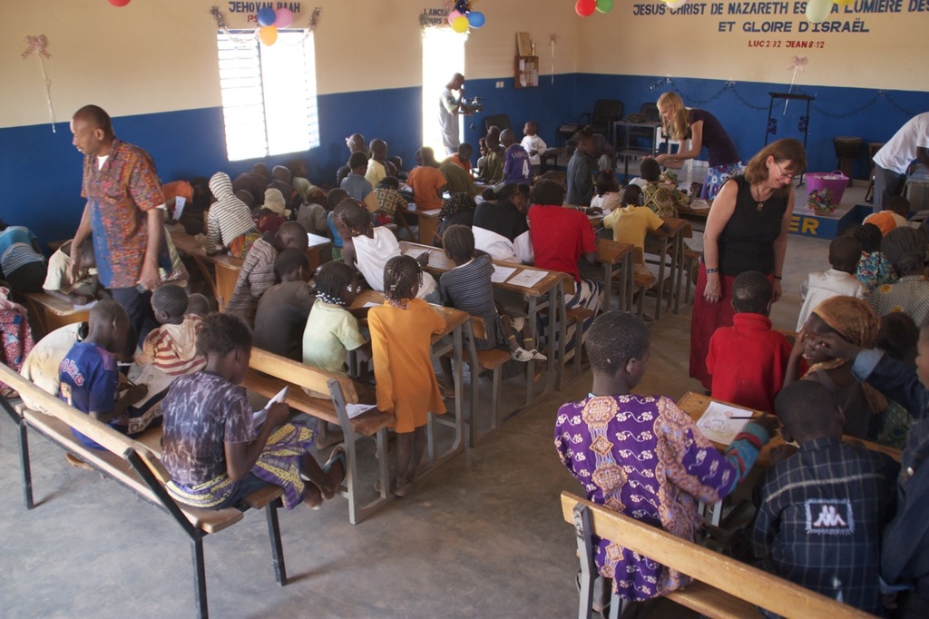 Kinder auf Bänken in Vordergrund mit Erwachsenen, die sie führen, Stühle, eine Tür, ein Fenster und ein Tisch im Hintergrund, Ballons und Text oben.