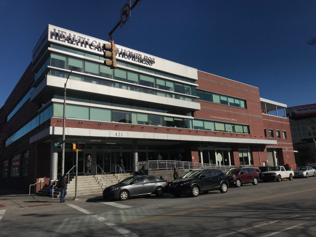 Ein großes Gebäude mit parkenden Autos davor, umgeben von Straßenlaternen, Verkehrspfosten, Verkehrsschildern, Menschen auf dem Gehweg, Geländern, Treppen, Bäumen und einem klaren blauen Himmel, identifiziert als das neue York City Health Care Homeless Center.