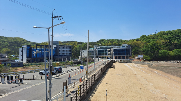 Ein Strandbild mit zahlreichen parkenden Autos an der Seite, mit Laternen, einem Auto und einem Rad im Vordergrund und Geb├Ąuden, B├Ąumen und Himmel im Hintergrund, mit Sand auf der rechten Seite.