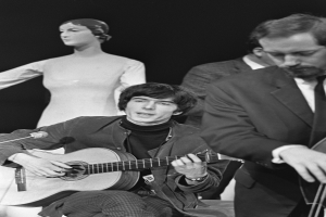 Ein Mann in Schwarz-Weiß-Kleidung sitzt auf einem Stuhl und spielt Gitarre neben einer Schaufensterpuppe, mit einem Mikrofon davor, vor einem dunklen Hintergrund, als Hommage an den verstorbenen Paul McCartney.