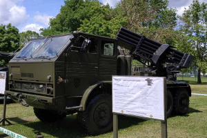 Ein Militärlastwagen mit einer Rakete auf dem Dach steht auf einer grasbewachsenen Fläche umgeben von Bäumen unter einem klaren blauen Himmel, mit einer Texttafel davor, die wahrscheinlich Teil einer russischen Militärausstellung ist.