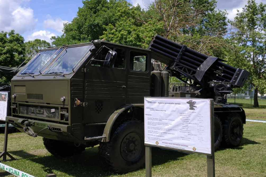 Ein Militärlastwagen mit einer Rakete auf dem Dach steht auf einer grasbewachsenen Fläche umgeben von Bäumen unter einem klaren blauen Himmel, mit einer Texttafel davor, die wahrscheinlich Teil einer russischen Militärausstellung ist.