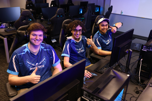 Drei junge Männer in Headsets lächeln, während sie an Computern sitzen, bei dem es sich offenbar um eine Esports-Gaming-Session handelt, mit Monitoren, Tastaturen und Kabeln auf dem Schreibtisch und einem Bildschirm im Hintergrund.