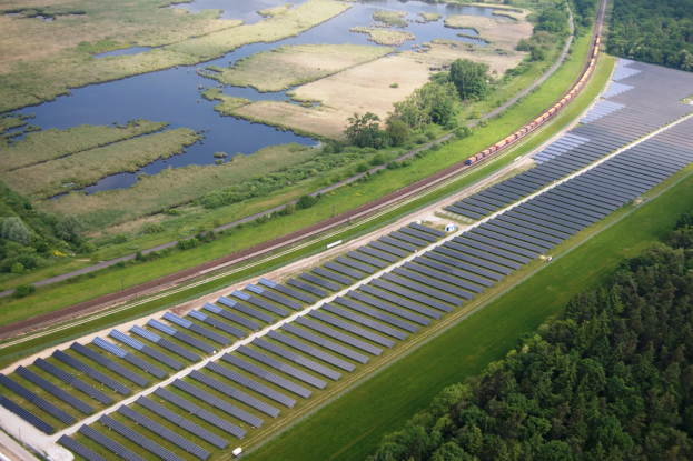 Luftaufnahme einer Solarpark mit Panelen in einem Feld, umgeben von Bäumen, Gras, Pflanzen und Wasser, mit einem Zug auf einer nähergelegenen Eisenbahnschiene.