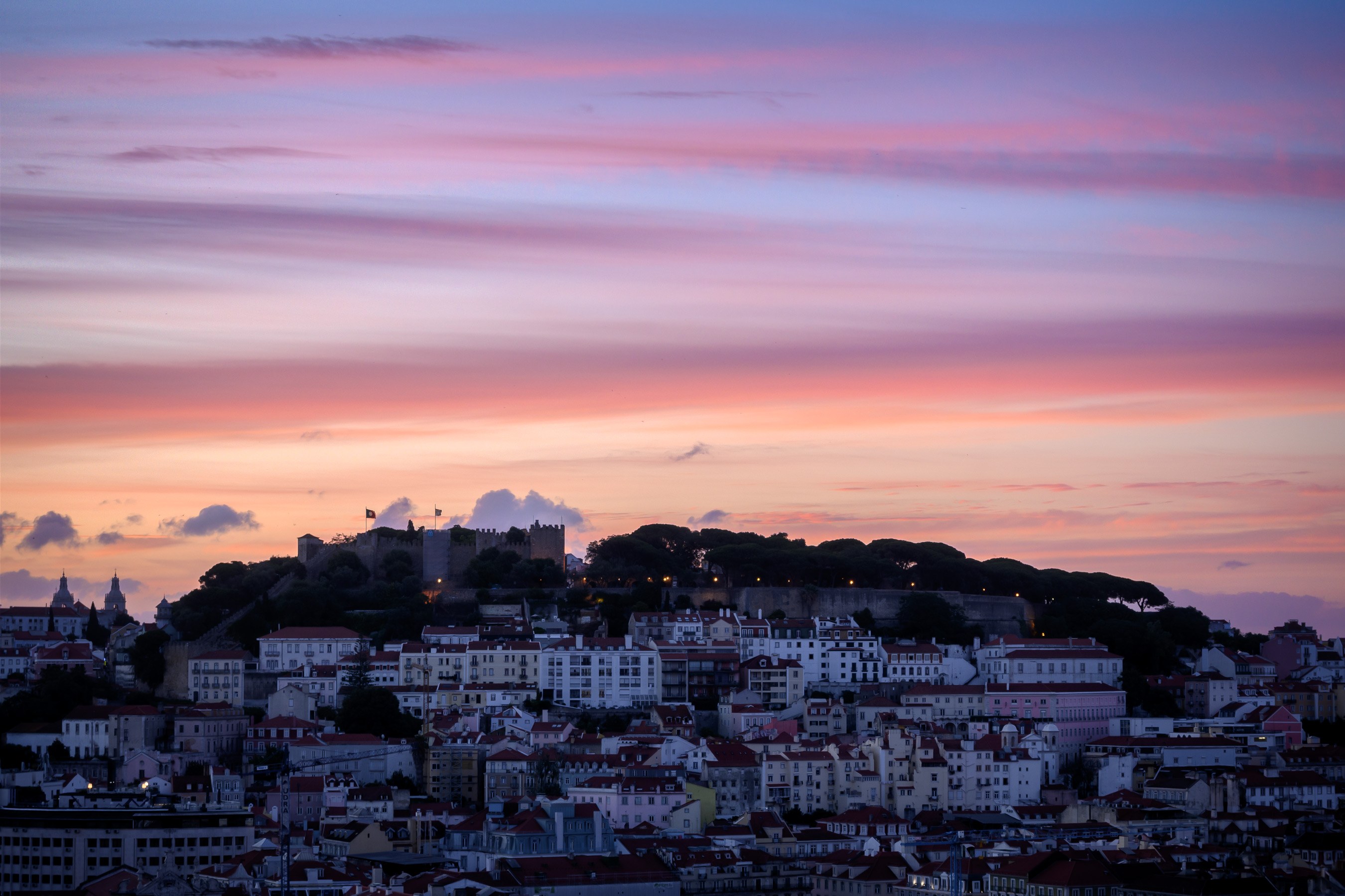 Sonnenuntergangsblick auf Lissabon, Portugal von einem Hügel aus, mit Gebäuden und Bäumen im Vordergrund und Wolken am Himmel.